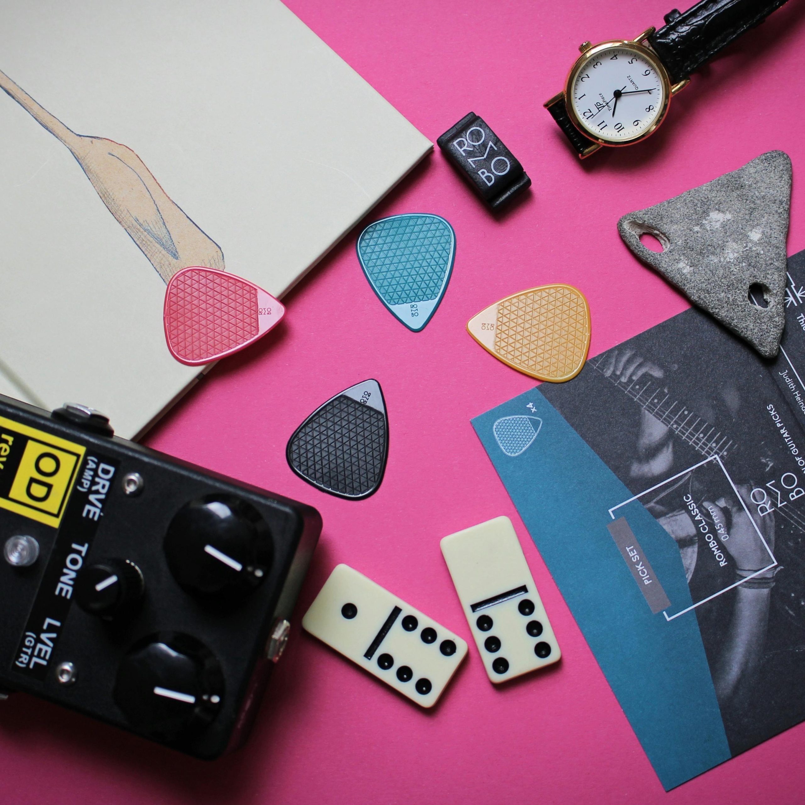 Guitar picks, dominoes, and a watch creatively arranged on a vibrant pink surface for a musician's aesthetic.